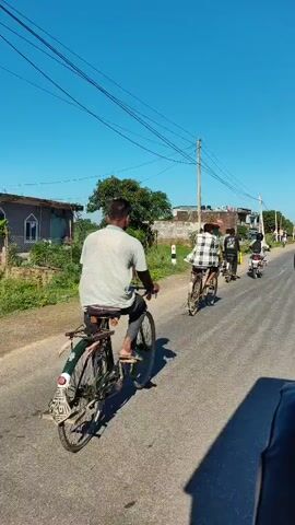 A video shows people riding bicycles and motorcycles on a paved road in a rural or semi-rural area. The sky is clear and blue, and there are trees and buildings visible in the background. The video appears to be filmed from a moving vehicle.