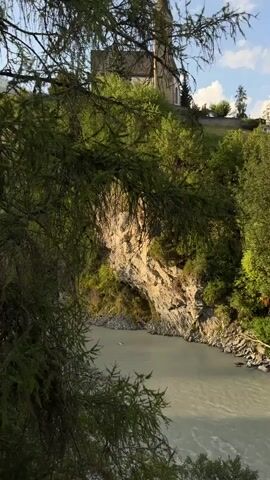 A still shot captures a natural landscape with a river flowing through a gorge. A rocky cliff face with vegetation rises from the riverbank. In the background, a church with a tall spire stands on a hill, surrounded by trees and a blue sky with clouds. The foreground is dominated by the branches of a coniferous tree, partially obscuring the view. There are no people or discernible actions in the video.