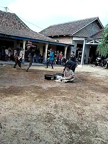 This video shows an outdoor event taking place in a rural setting. In the foreground, there is a dirt and mud-covered area, with some puddles of water. In the background, there are traditional houses with tiled roofs and open verandas. A group of people is gathered, seemingly watching a performance.

In the center of the scene, a person is sitting on the ground, tied up with ropes. Around them are two black buckets and some white material, possibly straw or fabric. Several individuals are actively involved in the event. One person, wearing a blue shirt and dark pants, is holding a whip or rope and appears to be directing or interacting with the person on the ground. Another individual, dressed in black, is standing near the seated person. Other people in dark clothing are also present, some holding what appear to be long sticks or whips.

The overall impression is of a cultural performance or ritual, possibly involving a form of traditional martial arts or a demonstration of skill and control. The audience is positioned along the verandas of the houses, observing the actions in the central area. The lighting suggests it is daytime, with overcast skies.