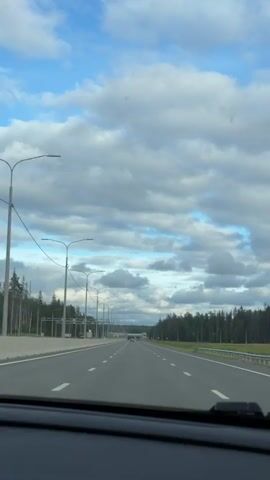 This video shows a first-person view from inside a car driving on a multi-lane highway. The road is paved with asphalt and has white lane markings. On the left side of the highway, there is a concrete barrier, followed by a grassy area with trees and streetlights. On the right side of the highway, there is a grassy area with trees. The sky is blue with scattered white clouds. In the distance, there are some structures, possibly toll booths or overpasses, and more trees lining the road. There are no other vehicles clearly visible in the immediate vicinity of the car. The overall scene depicts a typical highway driving experience on a cloudy day.