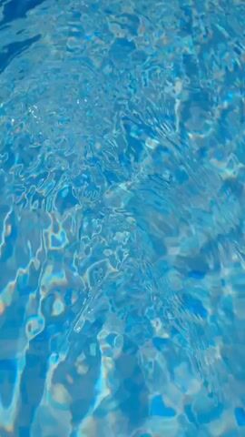 The video shows a close-up, vertical view of shimmering blue water. The water's surface is disturbed, creating ripples and waves that distort the light, causing it to refract and reflect in various shades of blue and hints of other colors. The texture of the water is dynamic, with areas of smooth flow and more turbulent patches where the light seems to be breaking. The overall impression is one of movement and light playing on a liquid surface, possibly a swimming pool or a body of water.