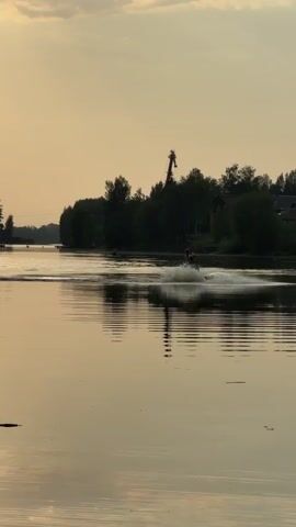 A person on a stand up personal watercraft is seen from a distance on a lake, at dusk. A person wears a yellow life jacket. The watercraft is a light color. The lake is generally calm and reflects the light of the sky. Some trees are visible along the bank, as well as a crane. A few birds fly overhead. A dock is visible in the distance.