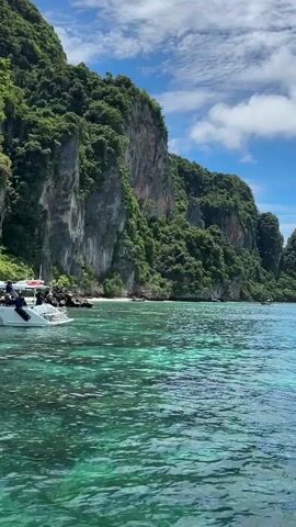 A boat with several people on board is moving through crystal clear turquoise water, close to a rocky, tree-covered cliff face. The boat is white with colorful graphics on the side, and the people appear to be enjoying the ride. The sky is blue with some white clouds.