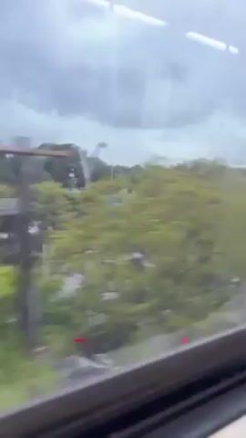 The camera is situated within a train carriage, viewing the outside world through a rain-streaked window. Outside, a multi-lane road bustles with vehicular traffic, including sedans, SUVs, and vans. Buildings, primarily white and modern in design, line the opposite side of the road. Trees, mostly green and lush, are interspersed along the roadside, and the sky above is heavily overcast with grey clouds. The scene suggests a rainy day in a suburban or urban environment, captured from a moving train.