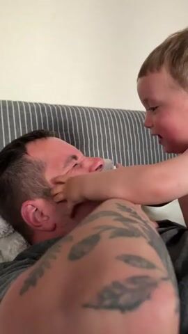 A man is lying on his back on a couch, facing upwards. A young child is leaning over him, smiling and touching his face. The man is smiling and appears to be tickled. The child's hand is touching the man's cheek. The child has light brown hair and is wearing a light-colored top. The man has short brown hair and is wearing a dark gray t-shirt. He has tattoos on his arm. The couch has a striped pattern and is against a white wall.