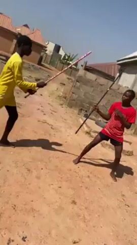 Two young, dark-skinned men are engaged in mock combat using sticks in an outdoor setting. The first man is wearing a yellow long-sleeved shirt and black shorts. The second man is wearing a red t-shirt with white polka dots and black shorts. They are both barefoot. Each man holds a long, slender stick, and they are positioned facing each other, simulating a sword fight with quick, deliberate movements. The ground is dirt and dry grass, with buildings and a blue sky in the background. The sun is shining, casting shadows of the figures on the ground.
