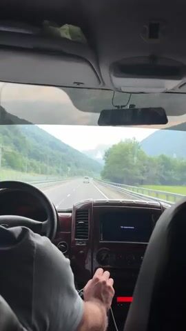 A first-person perspective video shows the interior of a vehicle moving along a highway. The driver's hands are visible on the steering wheel and gear shift. The dashboard features a navigation screen displaying "Нет видеосигнала" (No video signal). Outside, a winding road is bordered by lush green mountains, with a white van visible in the distance. The sky is overcast with clouds, and a faint mist hangs over the mountain peaks. The overall atmosphere suggests a scenic drive through a mountainous region.