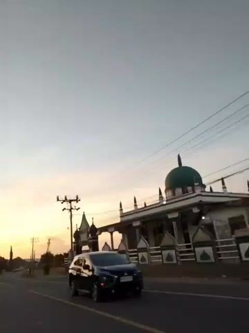 The video opens with a view of a mosque with a green dome and white accents, situated next to a road. An arched gateway with the word "MESJID" is visible. The scene is bathed in the soft light of a sunset, casting a warm glow across the sky, which transitions from orange to a pale blue. Overhead power lines crisscross the frame. The camera pans slightly, showing a road curving into the distance, flanked by palm trees and agricultural fields. There are several utility poles along the roadside. A motorcycle is partially visible on the left side of the road. The video continues to follow the road as it winds through the landscape, with the sunset providing a dramatic backdrop.