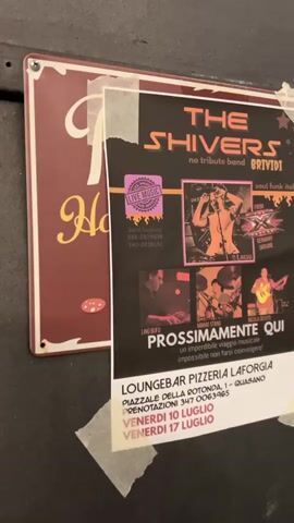 This is a close-up, slightly angled, handheld video of a poster advertising a live music event. The poster is dark with orange and pink text and graphics, and it is taped to a wall. Text on the poster reads "THE SHIVERS no tribute band" and "soul funk italiani" and "BRTVIDI". Below that, there are three smaller photos of musicians and text that reads "PROSSIMAMENTE QUI un imperdibile viaggio musicale impossibile non farsi coinvolgere!". At the bottom of the poster, the venue is listed as "LOUNGEBAR PIZZERIA LAFORGIA" with an address and phone number, followed by the dates "VENERDI 10 LUGLIO" and "VENERDI 17 LUGLIO". To the left of the poster is a partially visible maroon sign with yellow lettering that appears to say "Hot" with some cartoonish food images. The wall appears to be dark and textured. The lighting is dim.
