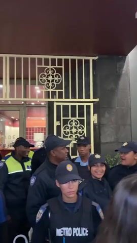The video shows a group of people walking indoors. The group contains a variety of individuals including security personnel in dark uniforms, one person in a bright yellow safety vest, and other casually dressed individuals. The environment includes a window with a view to the outside, architectural details in gold, and red or brown ceiling and wall sections. The movement is from right to left, suggesting the camera person is walking among the group, perhaps slightly ahead.