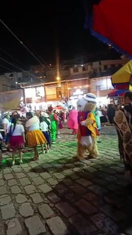 The video shows a nighttime street scene at a celebration. The street is paved with rectangular blocks and is crowded with people, most of whom are dressed in costumes. In the center of the frame is a large furry mascot wearing a white hat and a cape in the colors of the Ecuadorian flag. The mascot is walking among the crowd, which includes children in traditional dresses. In the background are buildings lit by streetlights, and a church is visible. A street vendor's colorful awning is partially visible in the upper right corner of the frame. The sounds of music and festivities are audible.