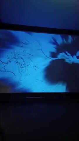 The video captures a large screen displaying dynamic, abstract visuals within a dimly lit indoor space, likely an exhibition or museum in Genoa, Italy.

Initially, the screen shows swirling blue patterns that transition to abstract green and cyan fluid-like movements. The display then shifts to a scene depicting a reddish-brown body of water with subtle ripples across its surface. A white boat, illuminated by lights, is visible on the right side of the screen. As the camera subtly pans, more of the reddish water and a dimly lit cityscape or port environment, featuring structures and lights, become visible in the background.

The visual progression suggests an immersive experience, potentially exploring themes related to water, maritime environments, or a port city's history and identity. Faint, indistinct text, appearing to be Italian words like "storia" (history) and "identità" (identity), briefly flashes on the screen.