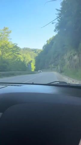 A dashcam view captures a winding road through a lush, green mountain landscape on a clear, sunny day. The road curves to the left, bordered by steep, tree-covered slopes on both sides. A metal guardrail runs along the right side of the road, separating it from the rocky incline. In the distance, a dark-colored car is visible, approaching the viewer's vehicle. The interior of a car is visible in the foreground, including the dashboard and windshield. The sky is a bright, cloudless blue.