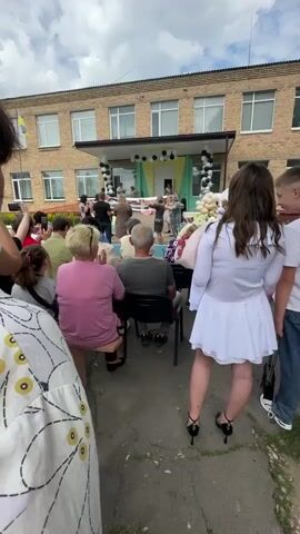 This media file captures an outdoor event taking place in front of a brick school building. The scene depicts a group of people gathered, with a stage set up at the entrance of the school. Decorations, including balloons and what appear to be banners, adorn the stage area. Several individuals are on the stage, seemingly participating in a performance or ceremony.

In the foreground, an audience is seated and standing, observing the activities on stage. The audience members include adults and some children. A young woman in a white dress and high heels stands facing away from the camera, holding a bouquet of balloons. To the left, a person in a white garment with a yellow circular pattern is partially visible.

The school building has multiple windows and a flag on a pole on the left side, suggesting a public or educational institution. The weather appears to be overcast with scattered clouds. The overall atmosphere seems festive, possibly a school celebration or graduation event, given the decorations and the presence of an audience. The time of day appears to be daytime, based on the lighting. No visible text is clearly legible in the provided frames. The location context is Horodyshche-Pustovarivske, Ukraine.