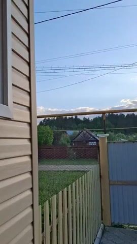 The camera pans from a light brown, wood-paneled building exterior to reveal a backyard scene with a gray metal fence, wooden fence, and grass. Beyond the fences are houses and a line of trees against a blue sky with clouds illuminated by the setting sun. Power lines stretch across the sky.