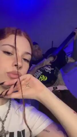 The video shows a young woman and a man. The woman is in the foreground, and the man is behind her playing a guitar. The woman has fair skin, a nose ring, long red hair styled in braids, and long black fingernails. She is wearing a white T-shirt, a silver chain, and a cross necklace. She is holding a strand of hair near her face, and she appears to be looking at the camera and pouting. The man has many tattoos on his torso and face, a shaved head, and is wearing a black t-shirt, a belt, and brown pants. He is playing a black acoustic guitar. The room is lit with a blue light, and there is a door with a window in the background.