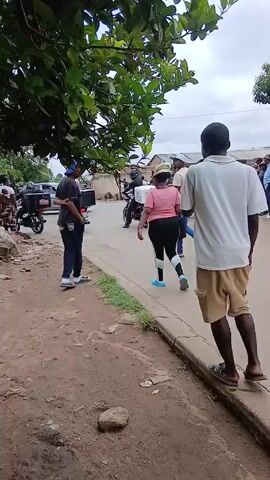 The video shows several people walking on or near the sidewalk along a paved road. Many appear to be walking in the same direction, although one man is walking away from the camera along the sidewalk. Other people are standing or walking around. Some are on motorcycles, one has a delivery box. There are also parked cars and buildings. The environment looks like a residential neighborhood. The sky is overcast.