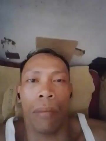 A close-up shot shows a person with short, dark hair, wearing a white tank top and black earbuds. The person is lying down, with pillows behind their head. The background appears to be a ceiling with some damage. The person’s face is the main focus, with their eyes open and looking forward.