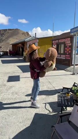 A person is captured outdoors in a sunny, mountainous landscape in Gyirong County, China. They are wearing a brown hat, a maroon jacket, blue jeans, and white sneakers, holding a large brown teddy bear dressed in a light-colored, possibly cream, outfit. The person is gently swaying or dancing with the teddy bear, facing away from the camera.

The scene is bathed in bright sunlight, casting clear shadows on the paved ground. In the background, simple, single-story buildings with various facades (yellow, brown) suggest a small village or a roadside rest stop. Beyond the buildings, barren mountains rise under a clear blue sky dotted with a few white clouds.

To the right, a tall signpost features a sign displaying "我在这儿枯措等你" (I'm waiting for you here at Kusu) and the number "4590," likely indicating altitude. Another smaller sign on a pole reads "48." and some Chinese characters. A building in the background has a sign with "石棺红色驿站" (Stone Coffin Red Post Station) and "我在这里等你" (I'm waiting for you here), accompanied by heart graphics, suggesting a scenic or romantic spot. In the right foreground, an outdoor table with a cup and potted plant, along with folding chairs, is visible. The atmosphere appears playful and relaxed.