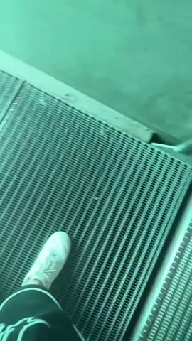 The video begins with a close-up of a person's feet wearing white sneakers and dark shorts with white writing on them, standing on a grated surface. The camera then tilts up to reveal a clear, turquoise body of water with numerous small fish swimming. The view appears to be through a glass pane, possibly a window or a transparent floor. The water is calm, and the lighting is bright, creating an underwater scene.