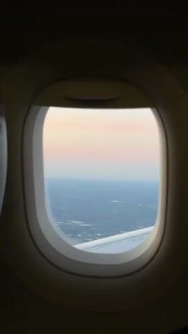 The video displays an interior view from an airplane window, capturing the world outside during what appears to be either sunrise or sunset. The oval-shaped window frame is prominent in the foreground, framing the aerial landscape.

Through the window, a vast expanse of land unfolds beneath a subtly colored sky. The sky transitions from soft pastel pinks and oranges at the horizon to lighter blues higher up, characteristic of twilight hours. Below, the ground is visible, appearing as a sprawling network of urban or suburban developments, with scattered lights or structures indicating human settlement across a relatively flat terrain.

A section of the airplane's white wing is visible in the lower right portion of the window, extending into the frame and providing a sense of the aircraft's movement and elevation. The overall scene is serene, offering a high-altitude perspective of the Earth under a fading or emerging light. There is no visible text in the video.