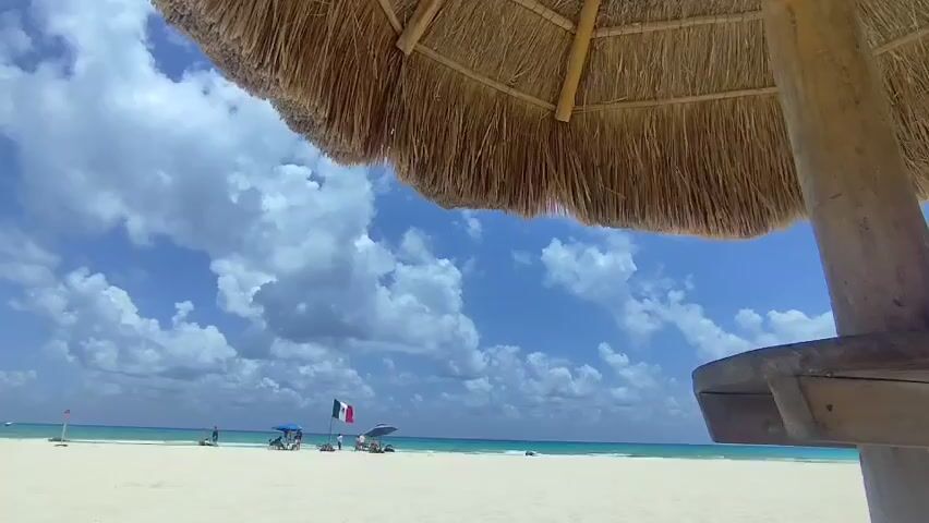 The camera is positioned beneath a thatched-roof beach umbrella, looking out at a sandy beach under a partly cloudy sky. The sand is light colored and appears relatively clean. In the distance, the turquoise ocean meets the horizon. Several people are scattered along the beach, some near umbrellas and others walking along the shore. Notably, a flag resembling the French flag (blue, white, red vertical stripes) is visible in the distance. The umbrella's wooden post is visible on the right side of the frame.