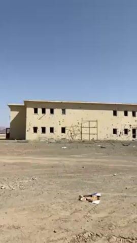 The video shows a two-story building that has been damaged, with numerous bullet holes and some structural damage. A burnt-out car wreck sits near the building. Several people are walking in front of the building, some of whom appear to be armed. The environment is arid and dusty. The sky is clear and blue.