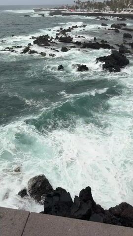 The video pans across a rocky coastline with waves crashing against the shore. The water is a turbulent mix of deep green and white foam, indicating rough conditions. Dark, jagged rocks jut out from the sea and line the shore, some partially submerged by the incoming waves. In the distance, a town or village is visible with buildings and some people walking along a pathway that runs parallel to the coast. The sky appears overcast, contributing to the somewhat dramatic and wild atmosphere of the scene. The camera starts from a higher vantage point looking down at the water and rocks, then slowly pans to the right, revealing more of the coastline and the distant settlement.