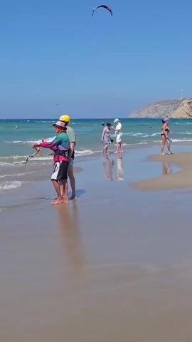This is a vertical video of a beach on a sunny day. The sand is wet near the waterline, creating reflections. In the foreground, two people are standing near the water, one holding a kite handle. A kite is visible in the sky. In the midground, several other people are walking on the beach or in the shallow water. Some are wearing swimwear, and one person is carrying a surfboard. In the background, there is the ocean with waves and a hilly coastline with wind turbines. The sky is clear and blue.
