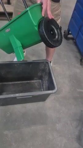 A person wearing a tan t-shirt, tan cargo shorts, and white sneakers is shown holding a green wheeled spreader over a black plastic tub. The spreader has two black handles and a black wheel. The tub is sitting on a gray concrete floor. In the background, there is a blue metal toolbox with wheels and a silver metal tool. The person is in the process of tilting the spreader to empty its contents into the tub.