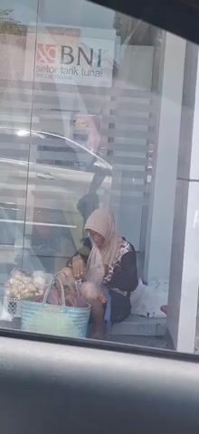 A person wearing a headscarf and a patterned shirt is sitting on a step outside a BNI bank. They are holding a plastic bottle and appear to be drinking from it. In front of them is a blue basket filled with items, and to their right, a white bag. The scene is viewed through a glass window, with reflections of a car and the sky visible.