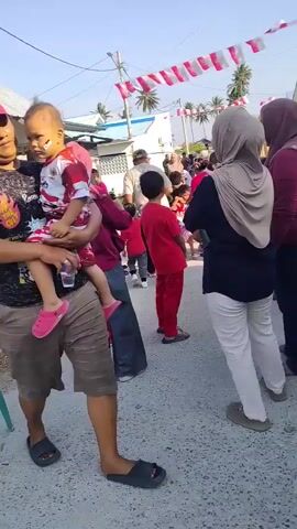 The video opens with a man holding a young child. The man is wearing a red baseball cap, a black t-shirt with a graphic print, and brown shorts. The child is wearing a red and white striped shirt and pink sandals. The man is holding a clear plastic bottle in his right hand. The scene appears to be outdoors, possibly at a street gathering or event, with a gravelly path and buildings in the background. Red and white decorative banners are strung across the scene. A group of people, many wearing red clothing, are visible in the background, some standing and some walking. There are also chairs and other objects scattered around. The camera then pans to the right, showing more of the gathering and the people present. The audio includes the sound of children's voices and a person speaking in Indonesian, mentioning that there are vendors selling food for those who are hungry or thirsty.