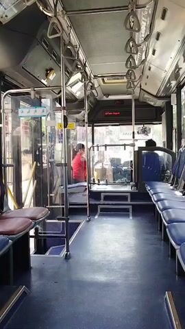 The video pans through the interior of a bus. The seats are blue and lined up along the sides. There are metal poles and handrails throughout the bus for passengers to hold onto. In the front section of the bus, a person in a red shirt is visible standing. The bus appears to be in motion, as indicated by the slight shake of the camera and the view through the windows, which shows trees and buildings passing by.