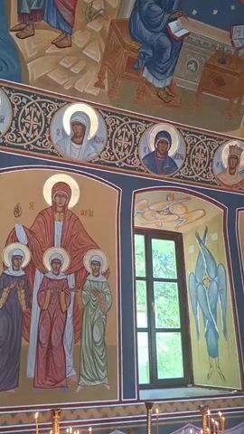 The video shows the interior of a church or monastery with religious artwork on the walls and ceiling. The artwork appears to be frescoes and icons depicting religious figures and scenes. The style of the artwork is consistent with Eastern Orthodox art. The room is decorated with a chandelier and there are people present in the lower part of the video. The environment is dimly lit, suggesting it may be evening or the interior is not brightly lit.