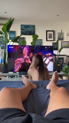 The video is a first-person perspective shot of two people in a bedroom. In the foreground, the legs and feet of the person filming are visible, lying on a dark blue comforter. To the right of the camera, a woman with long brown hair is sitting with her back to the camera, also on the bed, facing a large television screen. The television displays a live musical performance with a male singer on stage, accompanied by a band. The stage is lit with red and blue lights, and a large BBC logo is visible. The room is decorated with several framed pictures on the wall, including a landscape painting and nautical scenes. Two large potted plants are positioned on either side of the television. The lighting in the room appears to be natural, coming from the right side of the frame.