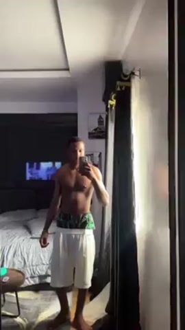 A shirtless man stands in a room, taking a selfie in a mirror. He is wearing white shorts with a colorful, patterned waistband. The room is decorated with a bed, a small round table, and a large black curtain with gold trim. A TV screen can be seen in the background.