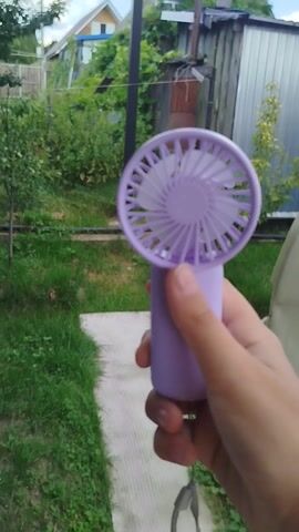 The video shows a close-up of a hand holding a small, light purple portable fan. The fan has a round grill with a patterned design and a cylindrical handle. A small grey string is attached to the end of the handle. The fan is being held horizontally, with the fan head to the left and the handle to the right.

The background is blurred but suggests an outdoor setting. Green grass and bushes are visible, as well as a section of a weathered wooden fence and what appears to be the side of a house with a white roof and windows. A metal structure, possibly a shed or industrial building, is also visible in the upper left background. The lighting suggests it is daytime, likely outdoors.

The hand is a person's, with visible fingernails. The person is holding the fan and appears to be interacting with it, possibly turning it on or off, as indicated by slight movements of the fingers around a button on the handle. The overall mood is casual and everyday. No text is visible in the frame. The location context provided is Oktyabrsky, Russia, which aligns with the visual cues of a rural or suburban outdoor environment.
