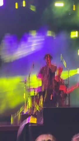 The video shows a live music performance. Two men are on stage. The man in the foreground is wearing a dark patterned shirt, black pants, and a hat. He is singing into a microphone and gesturing with his hands. He is standing on a platform or speaker. Behind him, another man is playing a guitar. A drum set is also visible behind the guitarist. Bright yellow and blue stage lights are illuminating the scene, with some smoke or haze present. A large screen is in the background with a circular graphic and text. The audience is visible at the bottom of the frame.
