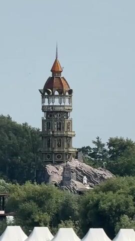 The video pans from right to left across a waterfront scene. In the background, a tall, ornate tower with a domed, copper-colored roof stands on a rocky outcrop amidst lush green trees. Below the tower, a line of white, tent-like structures are situated along the water's edge. Further to the left, a more modern building with glass elements is visible. In the foreground, the surface of a body of water is seen with gentle ripples. The sky is a clear, pale blue. The camera movement is smooth and steady, providing a panoramic view of the landscape. No people are clearly visible in the frame.