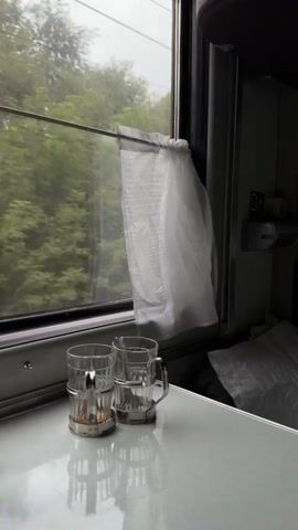 A video taken from inside a train compartment shows a close-up view of a small table by the window. On the table, two glass holders with glasses are visible. The glass on the left contains a small amount of liquid and possibly some tea leaves. The window shows a blurry view of green trees and a cloudy sky, indicating the train is moving. A thin white curtain is partially drawn over the window. In the background, bedding or pillows can be seen. The lighting is dim.