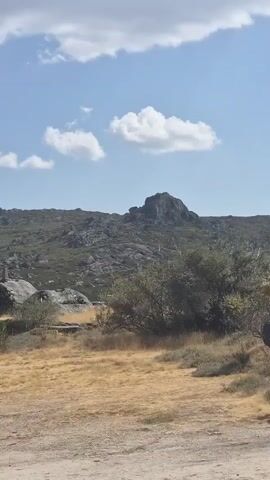 A wide shot shows a rocky, scrub-covered hillside under a bright blue sky with scattered white clouds. In the foreground, dry, golden grass and dirt cover the ground. A person walks from left to right across the scene, heading towards a body of water visible in the background.