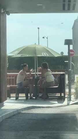 Two women are seated at a table under a green umbrella at an outdoor seating area of a fast-food restaurant. The woman on the left, wearing a white tank top and denim shorts, is holding a drink and facing the woman on the right. The woman on the right, wearing a yellow tank top and shorts, is facing away from the camera and towards the woman on the left. They appear to be in conversation. The table has some items on it, though they are not clearly discernible. The environment includes a wooden bench, a sidewalk, and greenery in the background, with a street lamp and a traffic sign visible. The lighting suggests it is daytime.