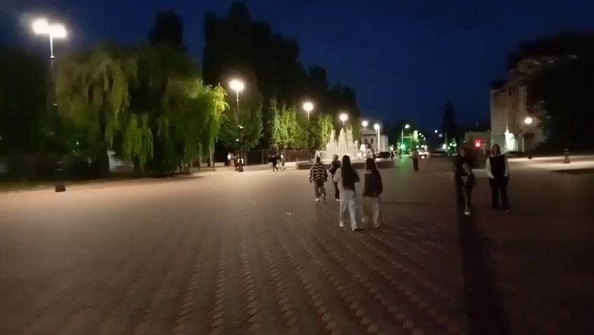 The video shows a paved plaza at dusk or night. Several streetlights illuminate the area, casting a warm glow. On the left side of the plaza, there are trees with weeping branches. In the background, a fountain is visible, with water spraying upwards. Several people are walking or cycling in the plaza. In the foreground, a group of three young women walks away from the camera. Further back, a group of people, some on bicycles, are also present. The overall atmosphere is relaxed and casual, suggesting a public space used for recreation and social gathering.