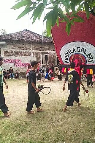 Here is a concise description of the video:

The video shows a group of people performing a traditional Indonesian dance or martial art form outside in a grassy area. Several individuals in black outfits wield whips as part of their performance. Behind them, a banner is visible, potentially identifying the group or event. In the background, there's a brick building and a crowd of onlookers. The video's vertical orientation suggests it was recorded on a mobile device.