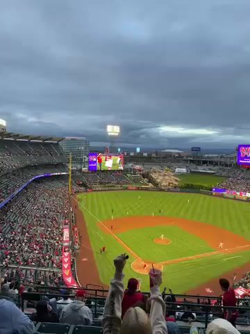 The video provides an elevated, wide-angle view of a baseball game in progress at a large stadium, likely Angel Stadium in Anaheim, United States. The setting is under an overcast sky, with stadium lights illuminated, suggesting either late afternoon or early evening.

Hundreds of spectators, many dressed in red, fill the grandstands, watching the game unfold on the green field with its red dirt infield and bases. Several baseball players are visible on the field. In the foreground, from the perspective of a spectator high in the stands, people are seated and engaged, with one individual in the center raising their arms in apparent excitement while holding a drink.

Large jumbotrons are prominent in the outfield, displaying graphics and possibly game information, though specific text is not clearly legible. The scene captures the lively atmosphere of a professional baseball game.