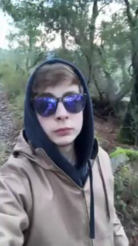 A fair-skinned person wearing mirrored sunglasses and a tan hooded jacket walks along a path in a forest.  The person has light brown hair and is wearing a dark-colored hoodie under the jacket. The trees in the background are blurry.