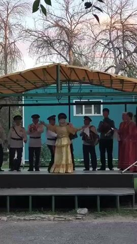 A group of people are performing on an outdoor stage. A man and a woman in traditional clothing are dancing in the center of the stage. Several men in military-style uniforms stand to the left of the dancers, some holding instruments or objects. To the right of the dancers, three men are playing accordions, and another man is holding a clarinet. A woman in a red dress stands on the far right. The stage is covered by a yellow awning, and a teal building is visible behind the performers. Trees and a cloudy sky form the background.