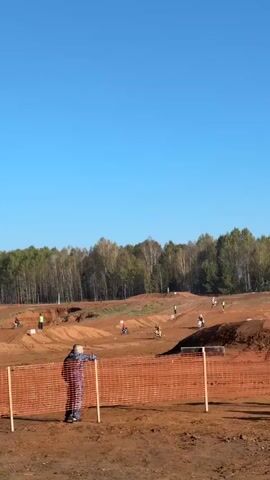 The video shows an outdoor motocross track on a sunny day. The track is made of dirt with several jumps and turns. Several people are on the track, riding motorcycles. In the foreground, a person stands behind an orange mesh fence, watching the race. Trees form a backdrop to the track.