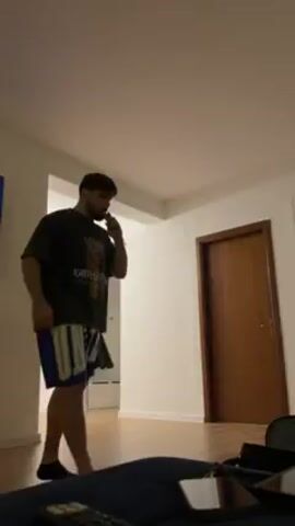 A man is dancing in an apartment. He is wearing a dark t-shirt and blue and white shorts. He has a beard and tattoos on his arms. He is holding something in his hand. There is a door behind him and a light fixture on the ceiling.  A bag and other items are on the floor in front of him.