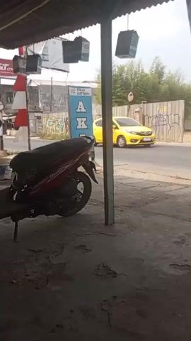 A video captures a street scene. In the foreground, the roof of an outdoor structure is visible, supported by pillars. A motorcycle is parked under this structure, partially obscuring the view of the street. Beyond the motorcycle, a street is visible with traffic. A yellow car drives by, followed by another vehicle. A red and white flag is seen mounted on a pole. A motorcycle with two riders passes by. The background shows buildings, fences, and some greenery. The overall lighting suggests it is daytime, possibly overcast.